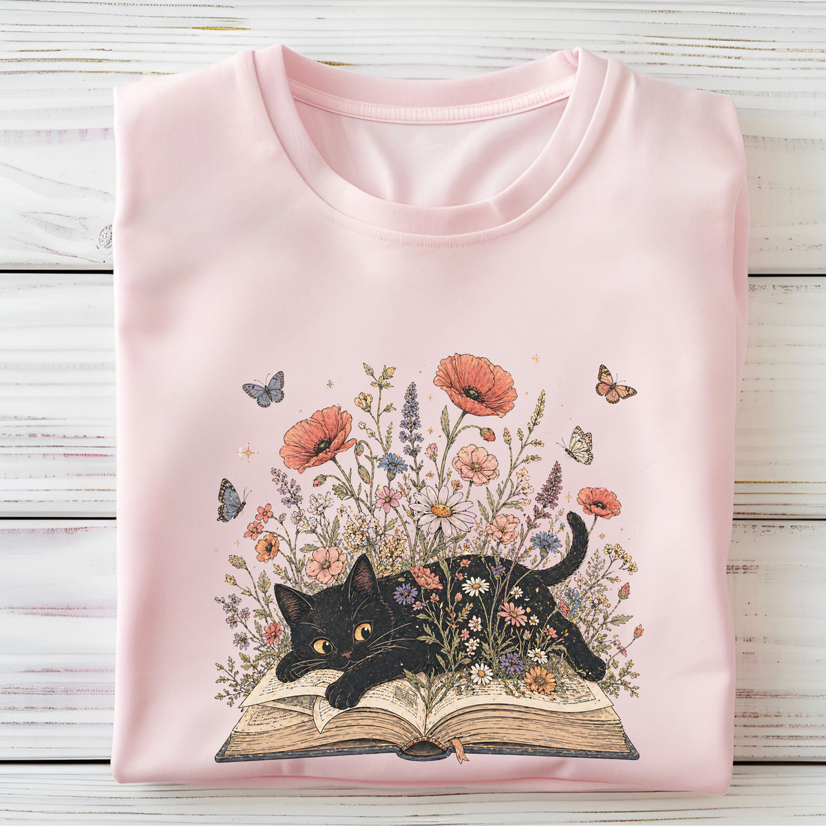A folded light pink crewneck t-shirt displayed flat on a white wooden plank background. The front of the shirt features a detailed graphic illustration of a black cat lying on an open book, surrounded by a vibrant garden of wildflowers and butterflies.