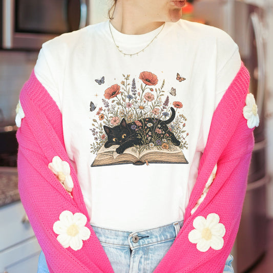 A person wearing a white graphic T-shirt with a black cat illustration over an open book and flowers, layered with a bright pink cardigan featuring white flower appliqués, paired with light blue jeans.
