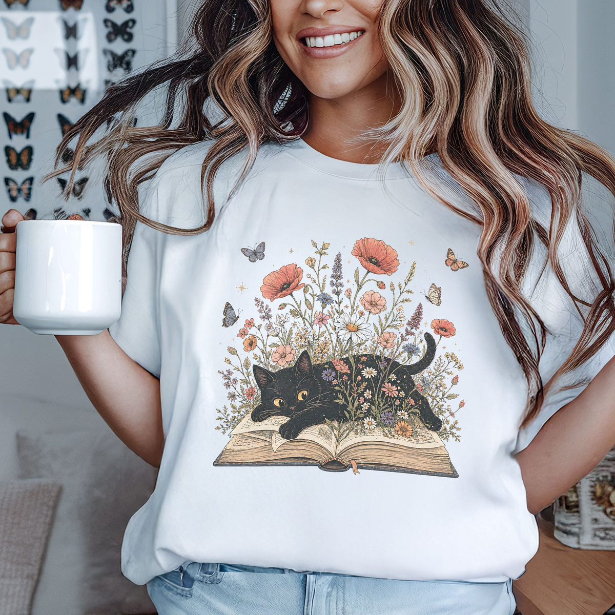 A woman wearing a white graphic t-shirt with a black cat emerging from an open book surrounded by wildflowers and butterflies, holding a white ceramic mug.