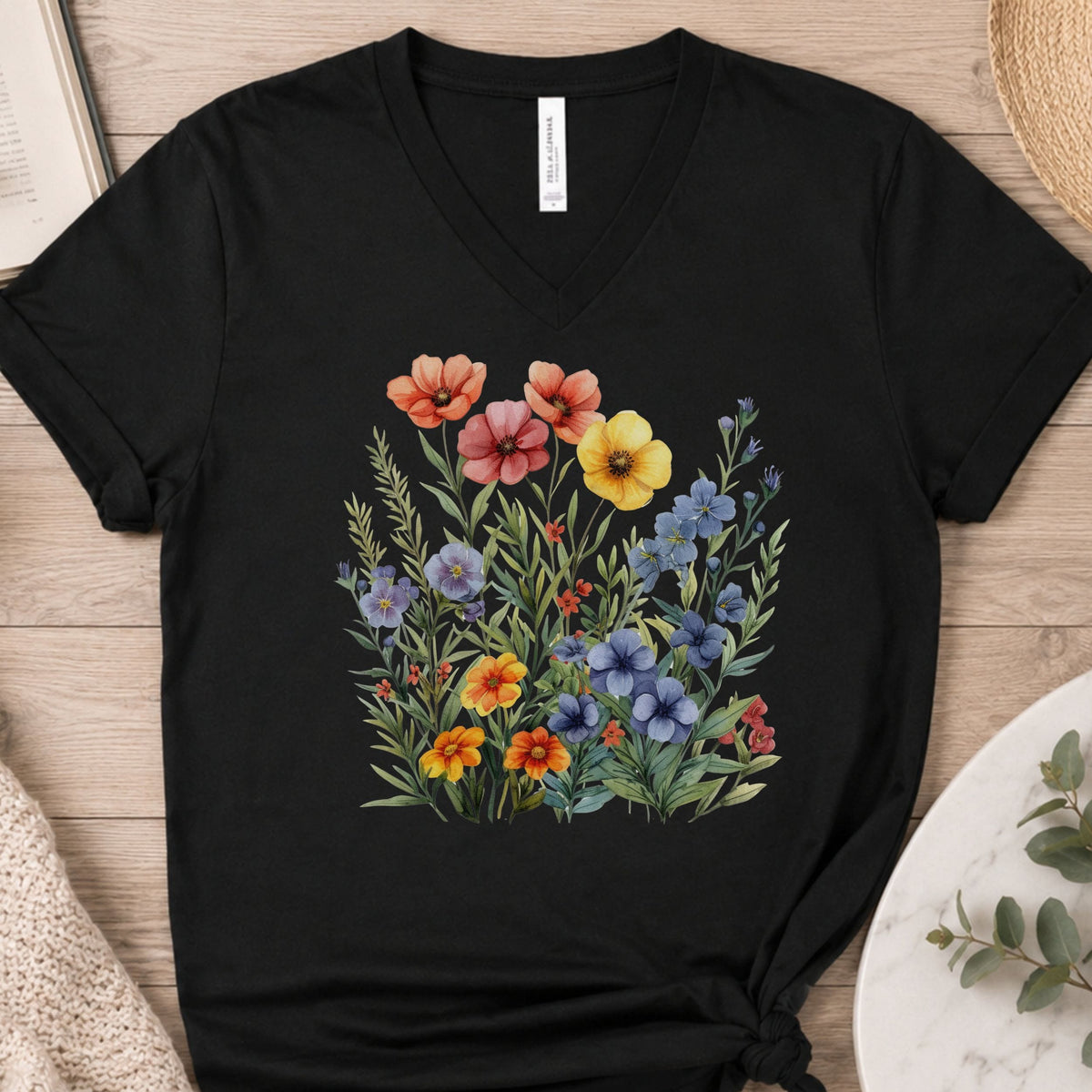 A black V-neck t-shirt with a colorful watercolor-style wildflower print on the front, laid flat on a light wood surface with styled props.