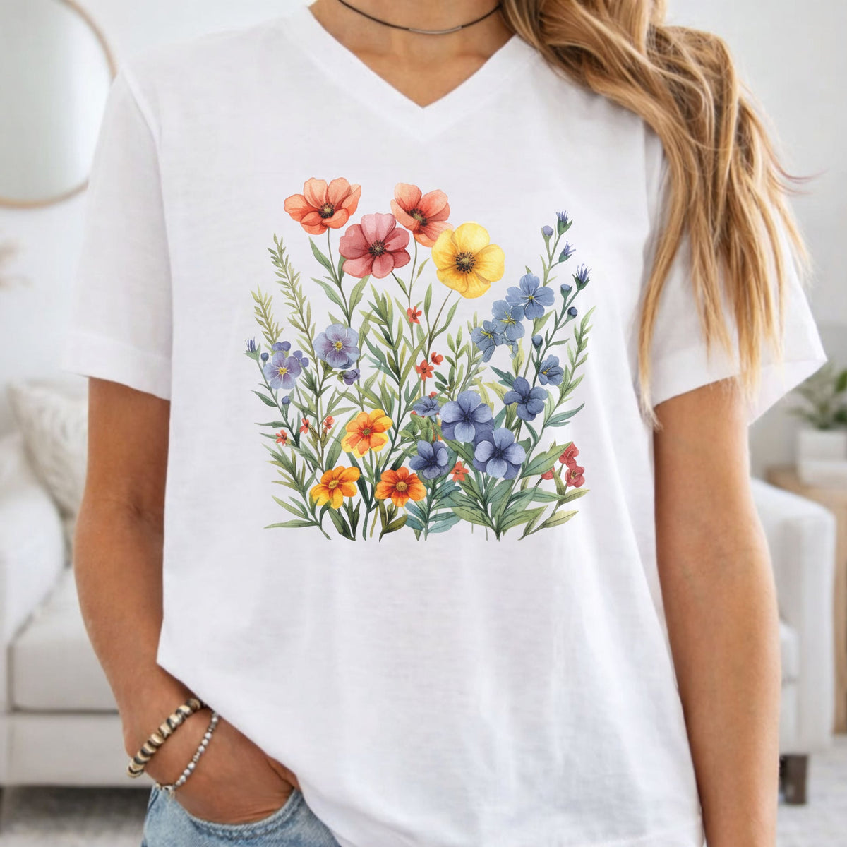 A woman wearing a white V-neck t-shirt with a colorful watercolor-style wildflower graphic on the front, set against a bright, softly blurred living room background.